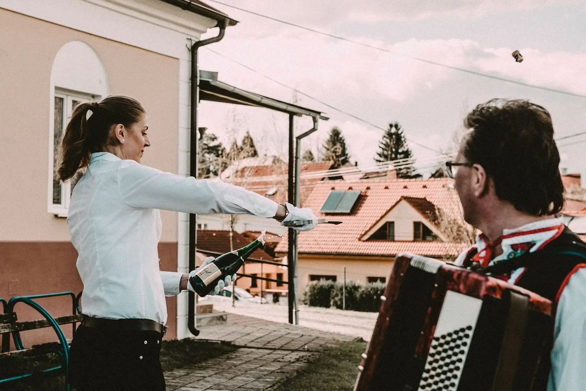 Obsluha otvára fľašu na terase, vedľa hrá harmonikár
