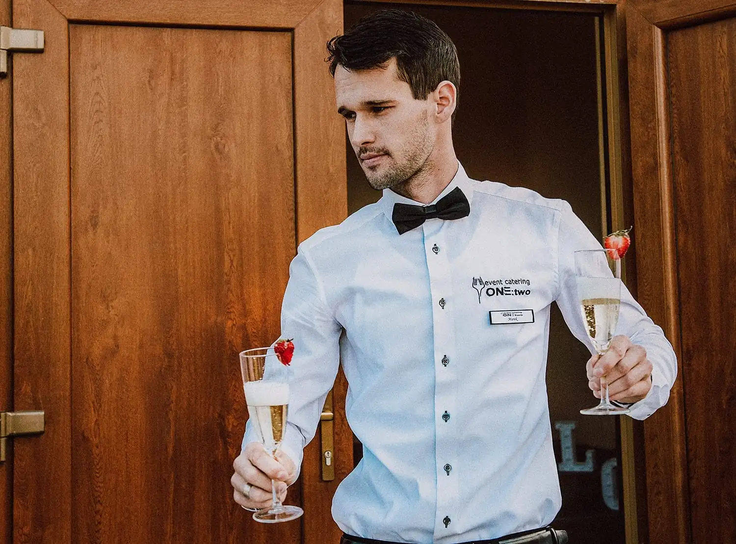 Čašník v košeli s logom ONE:TWO prináša dva poháre prosecca s jahodou